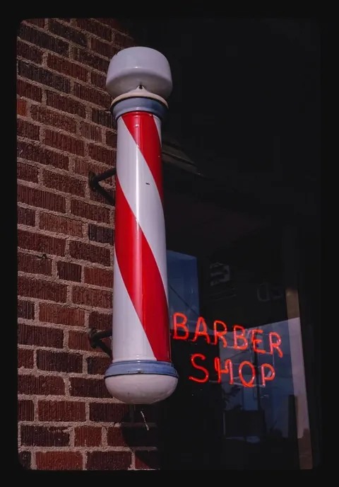 A red and white barber pole on a brick wall next to a neon Barber Shop sign